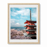 A framed art print featuring a Japanese  pagoda, pink cherry blossoms, mount Fuji, and a blue sky with clouds, displayed in a light wood frame against a white background.