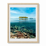 A framed split-level photograph of a tropical island above clear ocean water with a coral reef below, presented in a light wood frame with a white mat against a white background.