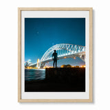 A framed photographic art print of the Sydney Harbour Bridge at night, featuring a silhouetted figure standing on a rock in the foreground. The print is matted with a white border and enclosed in a light natural wood frame, set against a clean white background.