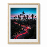 A framed vertical art print featuring a long-exposure photograph of a palm tree-lined Los Angles city street at dusk, with red light trails from cars, a blue and pink sky, and a white airplane contrail. The print is matted in white and enclosed in a light wood frame, shown against a plain white background.