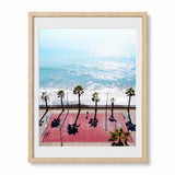 Framed wall art featuring an aerial photograph of a red basketball court at capistrano beach, with palm trees casting long shadows. Set in a light wood frame with a white mat.
