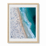 A framed aerial photograph of a bondi beach shoreline, showing a sandy beach crowded with small figures and turquoise ocean waves, presented in a light wood frame with a white mat against a white background.