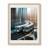 A framed art print depicting two silver subway trains in Chicago on elevated tracks between tall, reflective skyscrapers, presented in a light wood frame with a white mat against a light gray background.