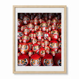 A framed photographic print of numerous red Japanese Daruma doll figures with white mats and a light natural wood frame, displayed against a clean white background.