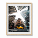 Framed wall art featuring a low-angle photograph of a city street with tall buildings converging toward the sky and a yellow taxi in the foreground, shown in a light wood frame with a white mat on a white background.