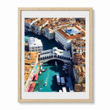 A framed aerial photograph of Venice's Grand Canal and Rialto Bridge, shown in a light wood frame with a white mat against a plain white background.