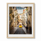 A framed art print depicting a scenic cobblestone street in Lisbon with a yellow tram, pedestrians, and colorful flags, presented in a light wood frame with a white mat against a white background.