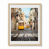 A framed photographic art print of a Lisbon street scene, featuring two yellow cable cars on a cobblestone hillside with buildings and tracks leading into the distance, presented in a light wood frame with a white mat against a white background.