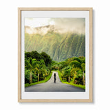A framed art print featuring a scenic photograph of a road in Hawaii lined with palm trees and lush green mountains shrouded in mist, presented in a light natural wood frame with a white mat.