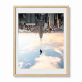 A framed surreal art print featuring a lone figure floating above clouds while a giant new york city skyline hangs upside down above them, presented in a light natural wood frame with a white mat against a white background.