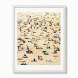 A framed art print depicting a crowded Bondi beach scene with numerous people sunbathing and walking across a sandy beach, presented in a white frame against a white background.