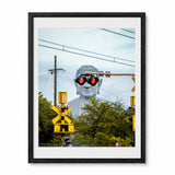 A Japan framed art print featuring a surreal photograph of a giant white Buddha statue wearing black sunglasses with red lenses, set against a backdrop of green trees, utility poles, and yellow railroad crossing signals. The print is presented in a white mat and a modern black frame against a clean white background.