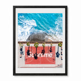A framed art print featuring an aerial photograph of a red basketball court with palm trees and the word 'Supreme' overlaid, at a California beach. The print is presented in a black frame with a white mat against a clean white background.