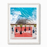 A framed art print featuring an aerial photograph of a red basketball court with palm trees and the word 'Supreme' overlaid, at a California beach. Displayed in a white frame against a light gray background.