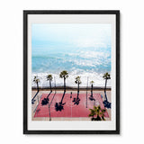 Framed wall art featuring an aerial photograph of a red basketball court at capistrano beach, with palm trees casting long shadows. The print is shown in a black frame with a white mat against a light gray background.
