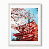 A framed art print featuring a red pagoda and pink cherry blossoms against a blue sky, presented in a white frame on a light gray background.
