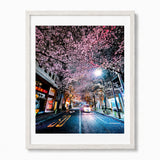 Framed wall art featuring a nighttime tokyo city street lined with blooming cherry blossom trees, shown in a light wood frame against a white background.