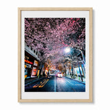 Framed wall art featuring a nighttime city street lined with blooming cherry blossom trees, shown in a light wood frame against a white background.