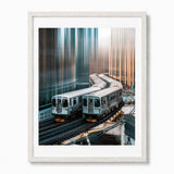 A framed art print depicting two silver subway trains in Chicago on elevated tracks between tall, reflective skyscrapers, presented in a white wood frame with a white mat against a light gray background.