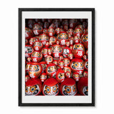 A framed photographic art print of a dense arrangement of traditional Japanese Daruma doll figurines, featuring a black frame and white mat board on a white background.