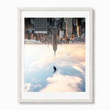 A framed art print featuring a surreal cityscape with new york city skyscrapers and a lone figure floating above a glowing blue and pink sky, presented in a white frame against a light gray background.