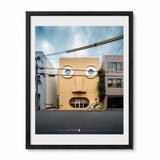 A framed art print featuring a whimsical photograph of face house in Kyoto, Japan - a yellow building with circular windows resembling a face, with a cyclist riding on the street in front. The print is framed in a black frame with a white mat against a white background.
