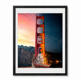 Framed wall art featuring a dramatic night and dusk view of the Golden Gate Bridge, with a warm orange-red bridge against a pink and blue sky, shown in a black frame with white mat on a light background.