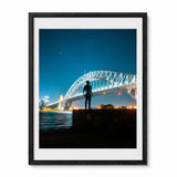 A framed photographic art print of Sydney Harbour at night, featuring a silhouetted figure standing on a rock in the foreground and the illuminated Harbour Bridge under a starry blue sky. The print is matted with a white border and enclosed in a black wood frame, shown against a plain white background.