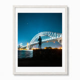 A framed photographic art print of the Sydney Harbour Bridge at night, featuring a silhouetted figure standing on a rock in the foreground under a starry blue sky. The print is housed in a white frame with a white mat, set against a clean white background.