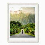 A framed art print featuring a scenic photograph of a road in Hawaii lined with palm trees and lush green mountains shrouded in mist, presented in a white woode frame with a white mat.