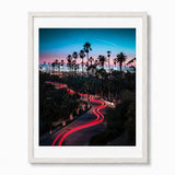 A framed vertical art print featuring a long-exposure photograph of a palm tree-lined Los Angles city street at dusk, with red light trails from cars, a blue and pink sky, and a white airplane contrail. The print is matted in white and enclosed in a white wood frame, shown against a plain white background.