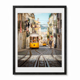 A framed photographic art print of a Lisbon street scene, featuring two yellow trams on cobblestone tracks between historic buildings. The print is presented in a black frame with a white mat board, shown against a plain white background.