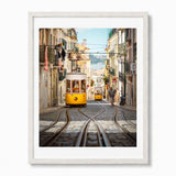 A framed photographic print of a Lisbon street scene featuring a yellow tram on a steep cobblestone hill, surrounded by historic buildings and a white frame against a light gray background.
