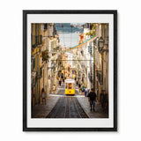 A framed photographic print of a Lisbon street scene with a yellow tram on cobblestone tracks between historic buildings, shown against a plain white background.