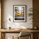 A framed vertical art print depicting a yellow tram in Lisbon, displayed on a beige wall above a wooden desk in a modern, sunlit room with a lamp, notebook, and chair.
