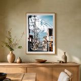 A framed art print depicting a scenic Japanese street with Mount Fuji in the background, displayed on a beige wall above a wooden console table with decorative vases and bowls.