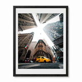 A framed photographic art print of a New York City street scene, featuring a yellow cab and surrounding skyscrapers viewed from a dramatic low angle. The print is framed in a black frame with a white mat, shown against a plain white background.
