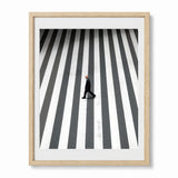Framed wall art featuring a high-angle photograph of a man walking across a black-and-white striped crosswalk, shown in a light wood frame with a white mat against a white background.