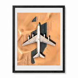 Framed art print featuring an aerial photograph of a white abandoned airplane sitting in a desert, with the black frame and white mat visible on a white background.
