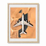 Framed art print featuring an aerial photograph of a white abandoned airplane sitting in a desert, with a natural wood frame and white mat visible on a white background.