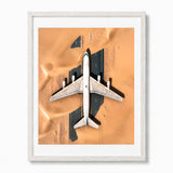 Framed art print featuring an aerial photograph of a white abandoned airplane sitting in a desert, with the white frame and white mat visible on a white background.
