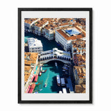 A framed aerial photograph of Venice's Rialto Bridge and Grand Canal, featuring turquoise water, white buildings, and terracotta roofs, presented in a black frame with a white mat against a light gray background.