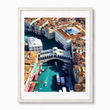 A framed art print of an aerial view of Venice's Grand Canal and Rialto Bridge, featuring turquoise water, historic buildings with terracotta roofs, and boats, presented in a white frame against a light gray background.
