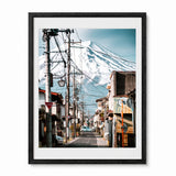 A black framed photographic art print of a Japanese street scene with a snow-covered mount fuji in the background, shown against a plain white wall.