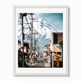 A framed photographic art print of Mount Fuji seen from a street in Japan, featuring a snowy mountain, utility poles, and buildings, presented in a white frame.