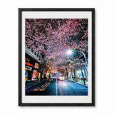 A black framed photographic art print of a Japanese city street at night, beautifully adorned with blooming pink cherry blossom trees over a wet road with streetlights and storefronts.