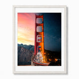 A framed photographic print of the Golden Gate Bridge, shown in a white frame against a light gray background. The print features a split view: the left side shows the bridge under a warm sunset sky with birds, and the right side shows the bridge under a starry night sky with a shooting star.