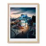 A framed photographic print of an izakaya in Tokyo at dusk, featuring a brightly lit building with blue signage, pedestrians, and a cloudy twilight sky. Set in a light wood frame.