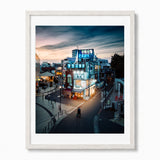 A framed photographic print of an izakaya in Tokyo at dusk, featuring a brightly lit building with blue signage, pedestrians, and a cloudy twilight sky. Set in a white wood frame