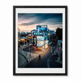 A framed photographic print of an izakaya in Tokyo at dusk, featuring a brightly lit building with blue signage, pedestrians, and a cloudy twilight sky. The print is matted with a white border and enclosed in a black frame, shown against a plain white background.